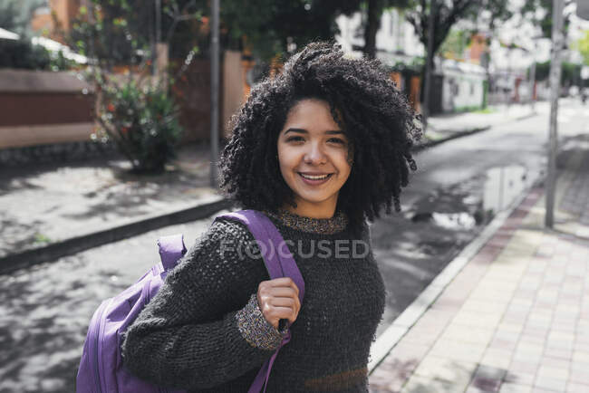 Side view of delighted ethnic female student with Afro hairstyle and backpack standing on street on sunny day and looking at camera - foto de stock