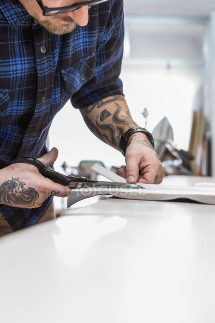 Recorte empanado pieza de corte artesanal masculino de tela en el banco de trabajo mientras se preparan materiales para la creación de tapicería de asiento de motocicleta en el taller - foto de stock
