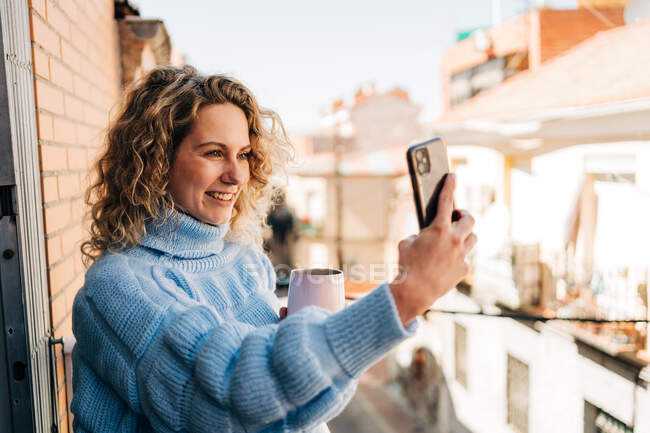 Vista lateral de la joven milenaria positiva con el pelo rubio rizado en suéter cálido elegante sonriendo mientras toma selfie en el teléfono móvil en el balcón a la luz del sol - foto de stock