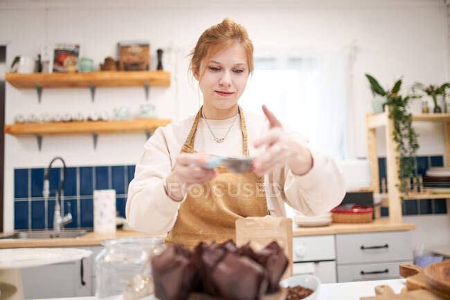 Sonriente hembra joven con tamiz rociando magdalenas en tazas de hornear con azúcar en polvo en la cocina de la casa - foto de stock