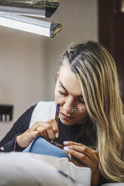 Cosmetologist with tweezers applying fake eyelashes for extension on eye of ethnic client with face protective mask in salon during coronavirus pandemic — Fotografia de Stock