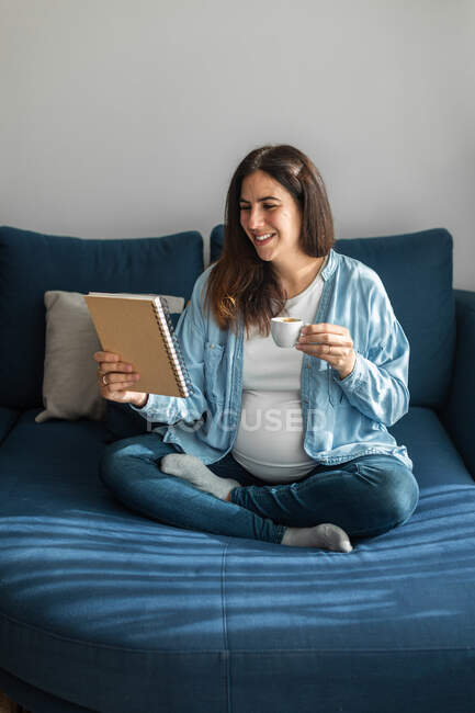 Mujer embarazada positiva sentada en un sofá blando con portátil y bebida caliente - foto de stock