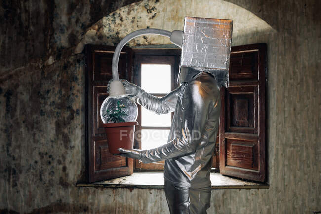 Вид сбоку неузнаваемый человек в серебряном костюме с дыхательным аппаратом и шлангом, прикрепленным к растению, стоящему в убогой заброшенной комнате — стоковое фото