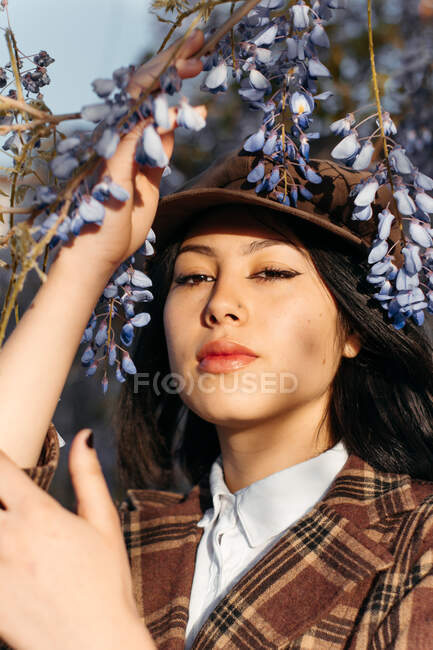 Young gentle ethnic female in checkered wear with beret touching blossoming plant while looking at camera on street — Fotografia de Stock