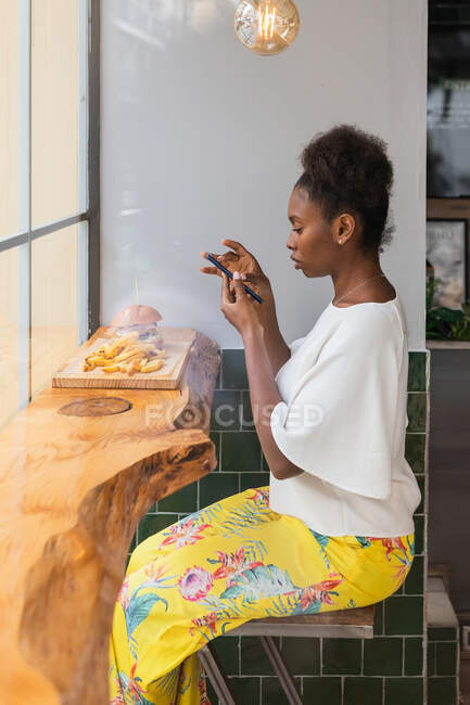 Seitenansicht ruhige Afroamerikanerin in stilvoller Kleidung, die Fotos von leckeren Burgern und Pommes frites macht, die auf einem hohen Tisch im Fast-Food-Restaurant serviert werden — Stockfoto