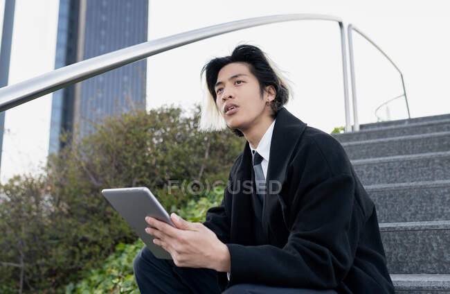 Young well dressed ethnic male executive browsing internet on tablet while sitting looking away on city staircase in daylight — Stock Photo