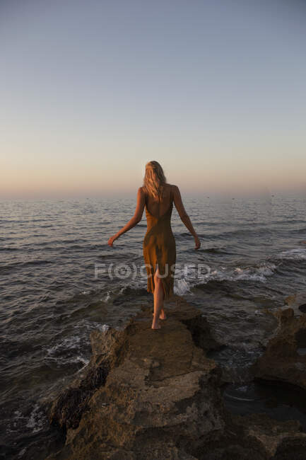 Back view of young woman walking on coast against blue waving sea at sunset — Stock Photo
