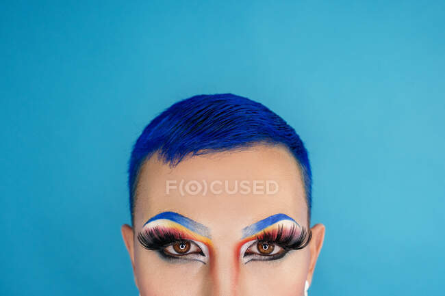 Crop queer with dyed blue hair and bright eye makeup standing on blue background and staring at camera — Photo de stock