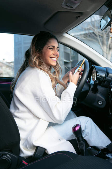 Side view of young content female in white apparel sitting in contemporary auto and looking away in city — Stock Photo