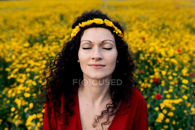 Сверху восторженная женщина в красном сарафане и с цветочной короной, стоящей с закрытыми глазами на цветущем поле с желтыми и красными цветами, наслаждаясь теплым летним днем — стоковое фото