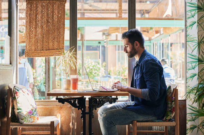 Seitenansicht eines männlichen Angestellten, der am Tisch mit leckerem Kuchenstück und Getränken an einem Netbook arbeitet — Stockfoto