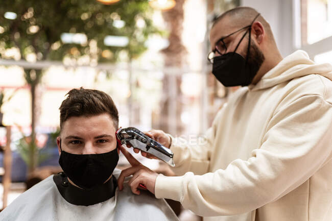 Peluquero masculino en anteojos haciendo corte de pelo al cliente adulto en peluquería durante la pandemia de COVID 19 - foto de stock