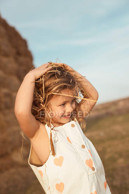 Alegre adorable niño en overoles jugando con heno cerca de pacas de paja en el campo - foto de stock