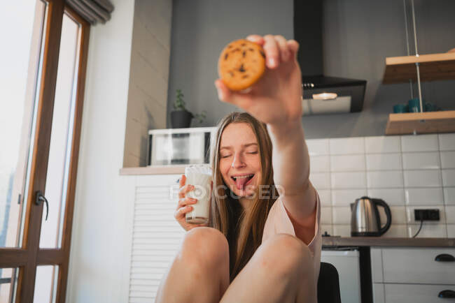 Молодая женщина с высунутым языком и закрытыми глазами сидит с овсяным печеньем и стаканом молока дома — стоковое фото