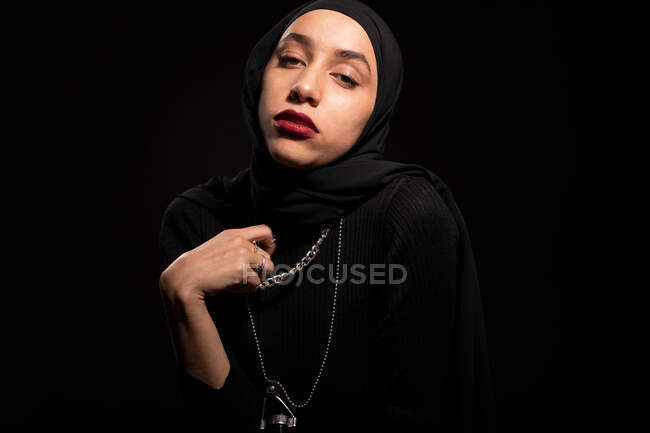 Jovem mulher islâmica atraente vestindo roupa preta e hijab gentilmente olhando para a câmera no estúdio preto — Fotografia de Stock