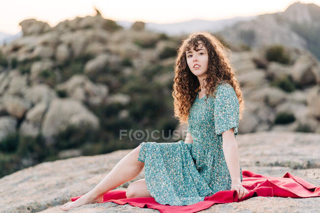 Young barefoot female in blue maxi sundress sitting on red plaid on rough rocky hilltop and looking at camera — Stock Photo