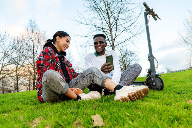 From below of cheerful multiracial couple in trendy wear with cellphone speaking on meadow with electric scooter in daytime — Stock Photo