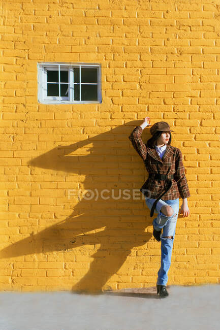 Stylische Frau in karierter Jacke und Baskenmütze, die wegschaut, während sie mit erhobenem Bein auf dem Bürgersteig gegen die gelbe Wand mit Schatten steht — Stockfoto