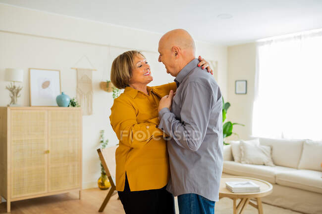 Счастливая пара средних лет обнимаются и танцуют в гостиной дома, глядя друг на друга с нежностью — стоковое фото