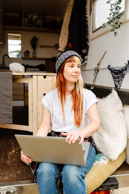 Delighted female traveler sitting outside on stairs of van while working remotely on laptop looking away — Stock Photo