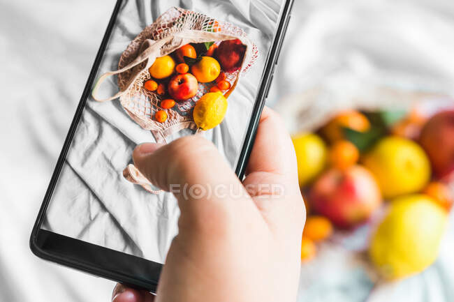 Зовнішній вигляд врожаю невпізнавана людина торкається екрана на мобільному телефоні, фотографуючи фрукти в мішку з нульовими відходами — стокове фото