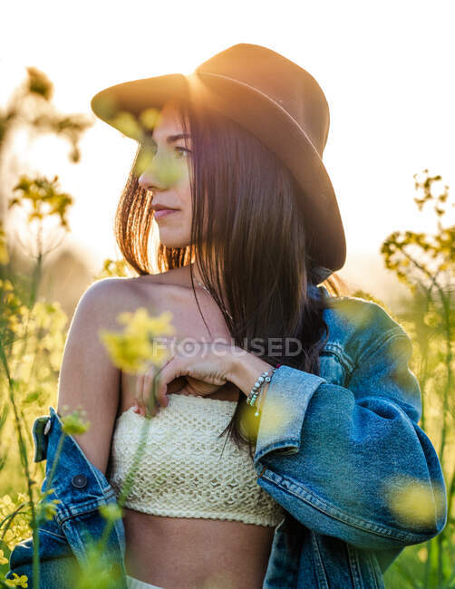 Молода брюнетка в білому верхньому капелюсі і нижній піджак стоїть, дивлячись на квітуче ріпакове поле в сонячний день — стокове фото