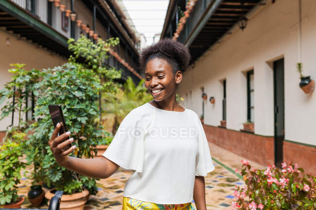 Felice giovane donna afroamericana indossa camicetta bianca prendendo selfie su smartphone mentre in piedi in cortile decorato con piante lussureggianti il giorno d'estate — Foto stock