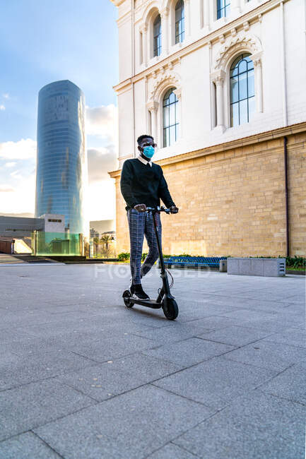 Unrecognizable ethnic male entrepreneur in sterile mask riding electric scooter on city walkway against buildings under cloudy blue sky — Stock Photo
