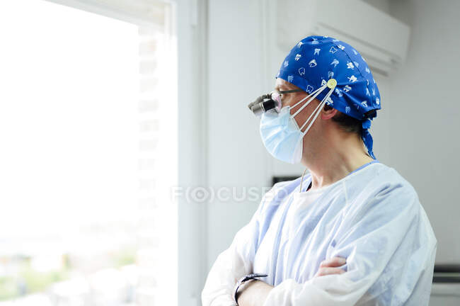 Анонімний лікар чоловічої статі в уніформі і стерильній масці, дивлячись подалі зі складеними руками проти вікна в лікарні — стокове фото