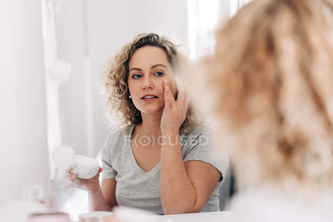 Содержание молодой женщины в повседневной рубашке, нанося увлажняющий крем на лицо и глядя в зеркало, сидя за столом тщеславия в светлой спальне — стоковое фото