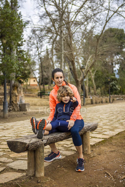 Charming boy pointing away while sitting in a wooden bench with mother in daylight in a park — Fotografia de Stock