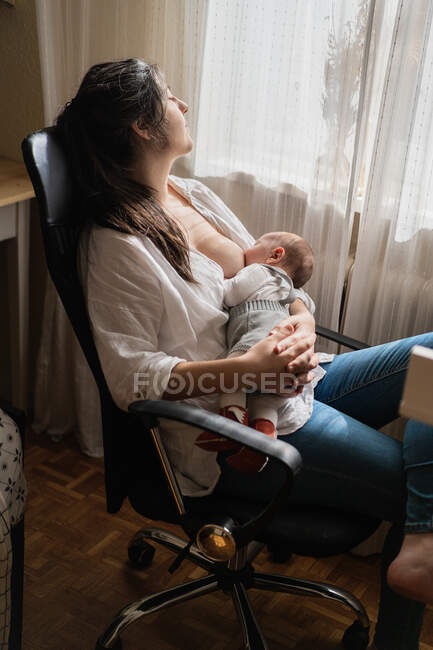 Maman adulte en tenue décontractée allaitant charmant petit enfant assis dans la pièce de la maison légère — Photo de stock