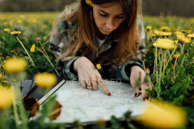 Concentrated female traveler with paper map and blossoming flowers looking away while lying on meadow against mountain in countryside - foto de stock