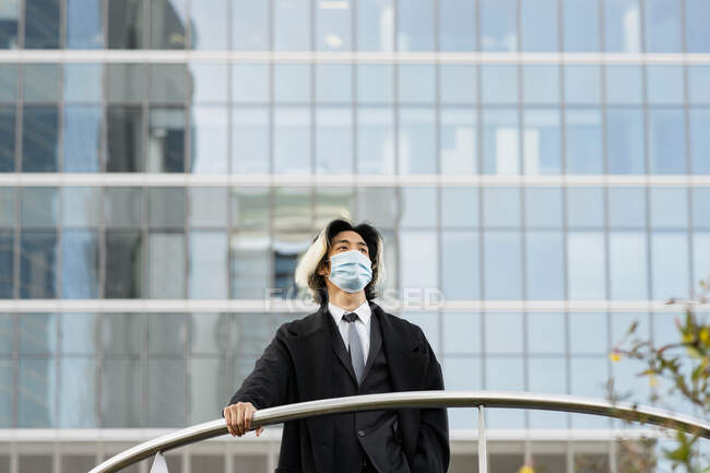 Молодий етнічний чоловік-підприємець у стерильній масці та пальто, торкаючись паркану, дивлячись на сучасний міський будинок — стокове фото