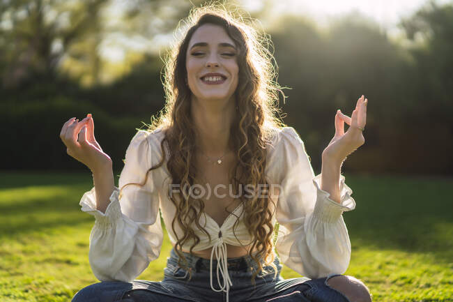 Contenida joven hembra con los ojos cerrados usando ropa casual sentada en Lotus Pose en verde claro hierba y meditando en el soleado parque - foto de stock