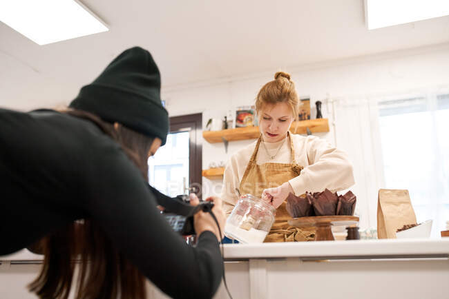 Crop photographe anonyme avec appareil photo contre blogueur avec pot de sucre glace et muffins dans des tasses à pâtisserie à la maison — Photo de stock
