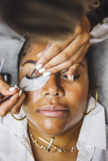 High angle of crop unrecognizable cosmetologist with tweezers applying fake eyelashes for extension on eye of ethnic client in salon — Fotografia de Stock