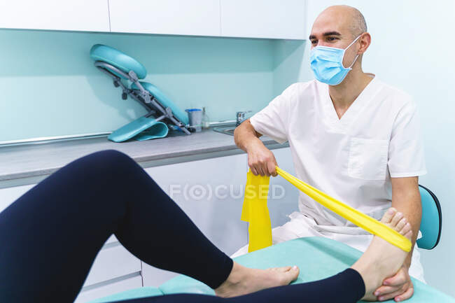 Male physiotherapist in medical mask with elastic band speaking with crop unrecognizable woman during rehabilitation process in hospital — Stock Photo