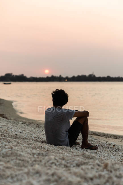 Вид ззаду на повну довжину невизначений чоловік у повсякденному літньому одязі відпочиває на піщаному узбережжі і насолоджується мальовничим видом на захід сонця — стокове фото