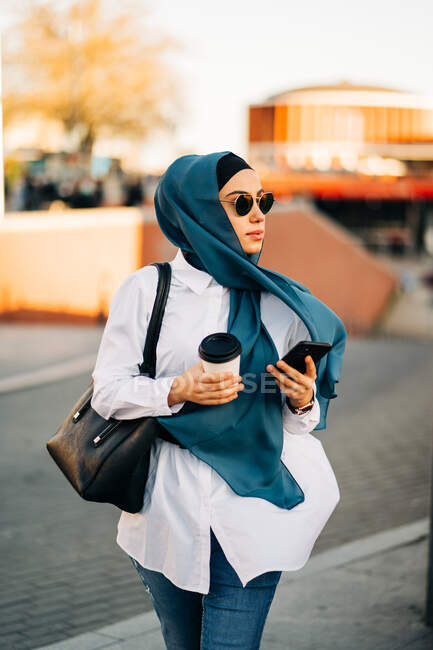 Ethnic female in headscarf and stylish sunglasses walking with takeaway drink on street and holding smartphone and looking away — Fotografia de Stock