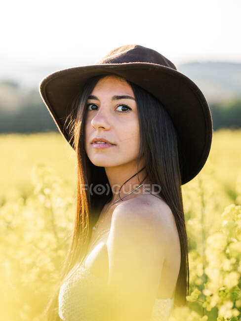 Вид збоку приваблива чутлива жінка з голими плечима і капелюхом, що стоїть серед ніжних жовтих квітів, що розквітають на просторому полі сонячної природи, дивлячись на камеру над плечем — стокове фото
