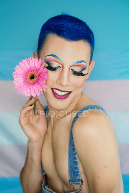 Joyeux drag queen avec un maquillage extravagant et des cheveux bleus touchant le visage avec une fleur rose tout en se tenant contre le drapeau transgenre — Photo de stock