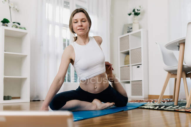 Faible angle de contenu flexible femelle assise dans la pose du roi Pigeon et regarder tutoriel en ligne sur tablette tout en pratiquant le yoga à la maison — Photo de stock
