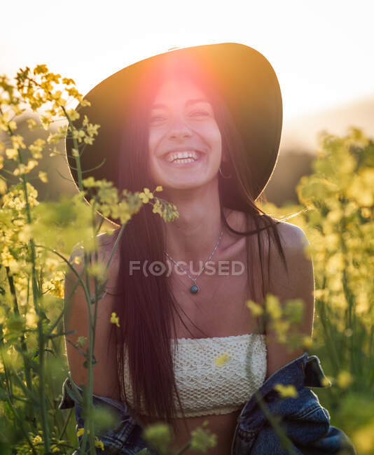 Delighted young brunette in white top hat and lowered jacket laughing merrily on blooming rapeseed field on sunny day — Stock Photo