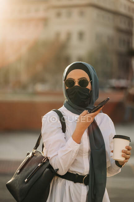Ethnic female in headscarf and stylish sunglasses standing with takeaway drink on street and recording voice message on mobile phone — Foto stock