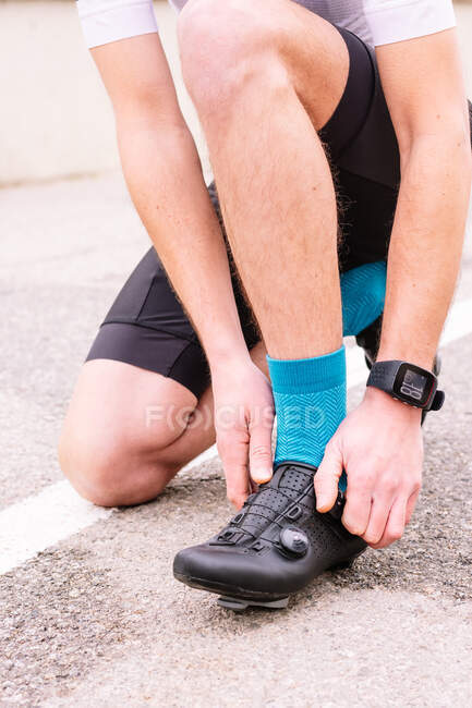 Crop unrecognizable male bicyclist in sports clothes and modern cycling shoes squatting on roadway against bike — Stock Photo