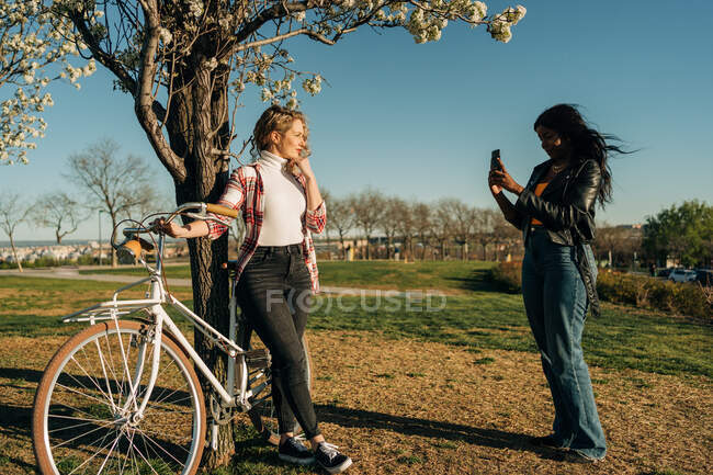Vue latérale de la femme noire prenant une photo d'une amie debout avec un vélo près d'un arbre dans un jardin de printemps — Photo de stock