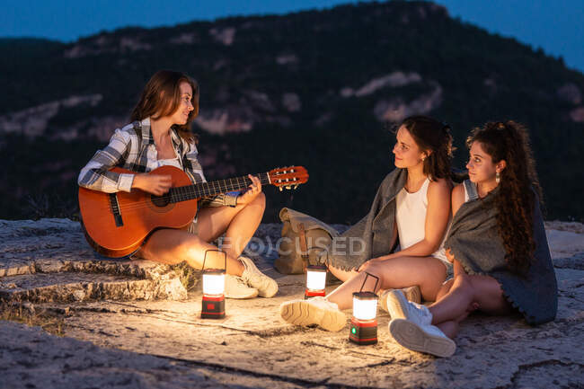 Side view of females travelers sitting on rock with glowing lantern and playing acoustic guitar while enjoying evening in highlands — Stock Photo