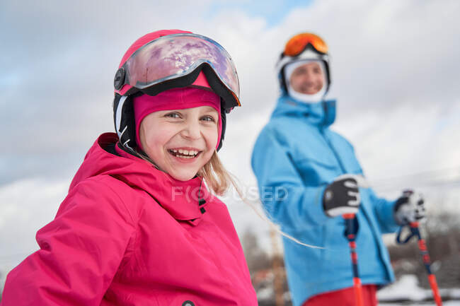 Menina alegre vestindo capacete de esqui e activewear de pé perto pai borrado em terreno de inverno nevado e olhando para a câmera com rosto sorridente — Fotografia de Stock