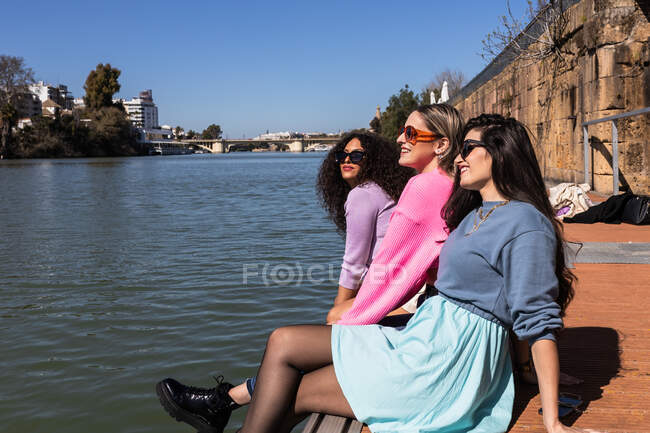 Alegres amigas multiétnicas em trajes casuais sentadas à beira-mar da cidade e olhando para longe com sorrisos no dia ensolarado de verão — Fotografia de Stock